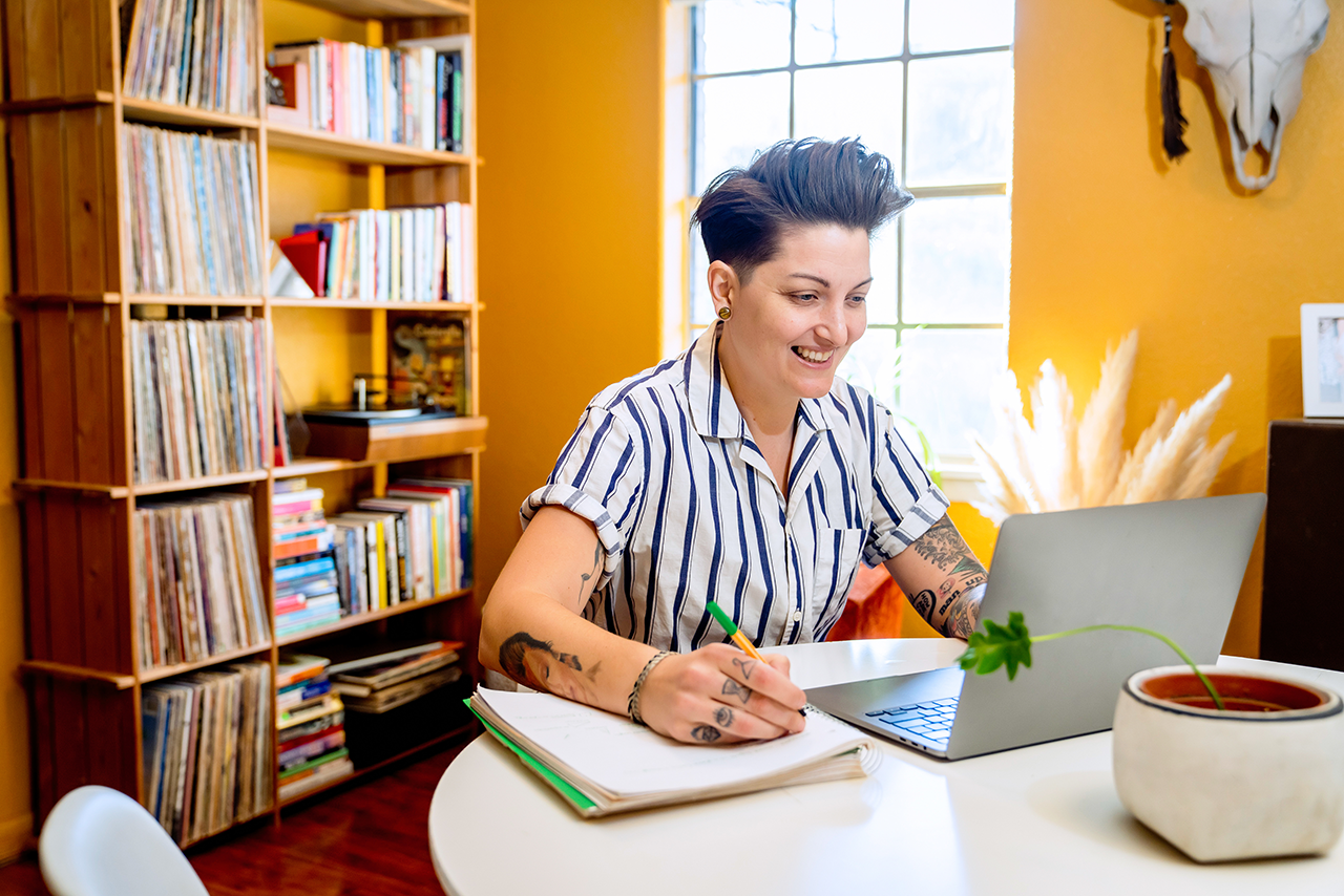 photo of gender queer person at a desk behind a computer taking notes on a notepad